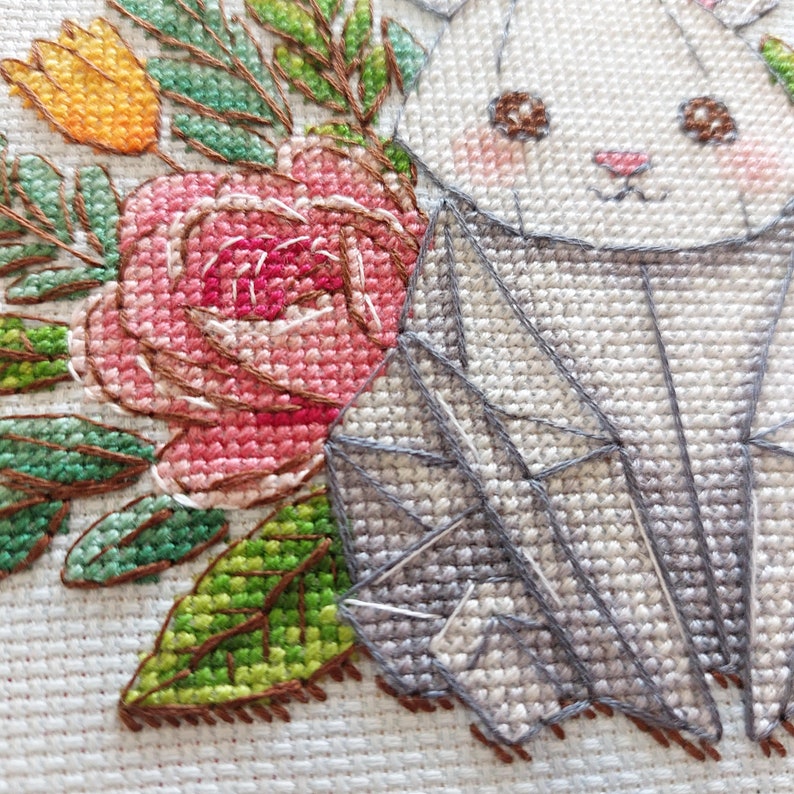 May include: A cross-stitch design featuring a white bunny with pink cheeks and a pink nose, surrounded by a pink rose, yellow tulip, and green leaves. The bunny has brown eyes and a geometric design on its body.