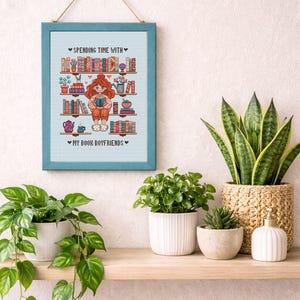 May include: A framed cross-stitch artwork with a teal frame, depicting a girl reading surrounded by bookshelves. The text reads "Spending Time With" and "My Book Boyfriends". The artwork is hanging above a wooden shelf with potted plants and a decorative basket.