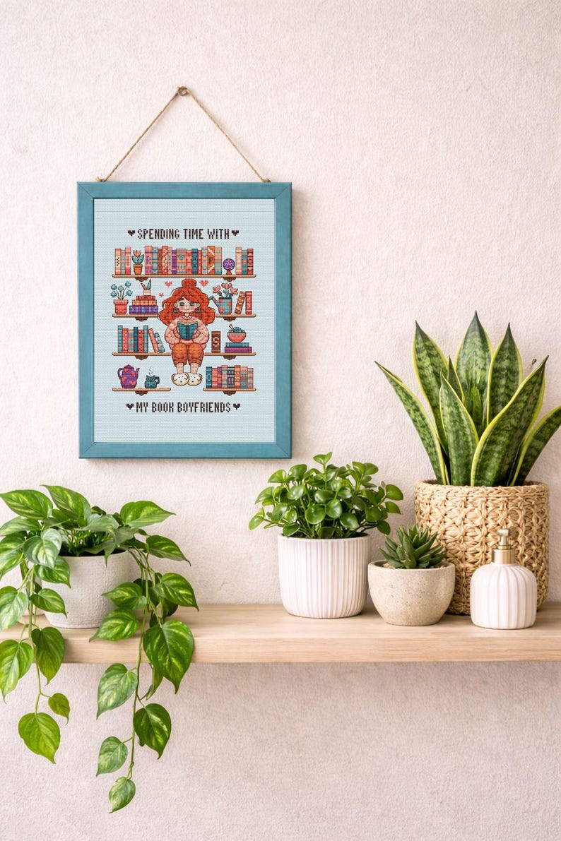May include: A framed cross-stitch artwork with a teal frame, depicting a girl reading surrounded by bookshelves. The text reads "Spending Time With" and "My Book Boyfriends". The artwork is hanging above a wooden shelf with potted plants and a decorative basket.