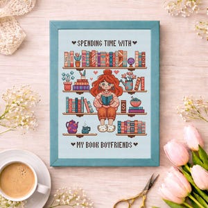 May include: Cross-stitch art framed in a light blue frame. The artwork depicts a person reading a book in front of a bookshelf. The text reads "Spending Time With" and "My Book Boyfriends". A cup of coffee, scissors, and flowers surround the frame.