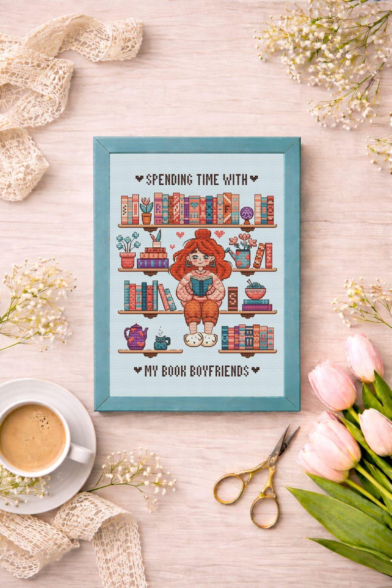 May include: Cross-stitch art framed in a light blue frame. The artwork depicts a person reading a book in front of a bookshelf. The text reads "Spending Time With" and "My Book Boyfriends". A cup of coffee, scissors, and flowers surround the frame.