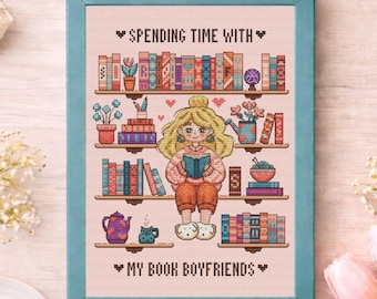 May include: A framed cross-stitch artwork featuring a cartoon woman reading a book, surrounded by bookshelves filled with colorful books and plants. The text reads "Spending Time With" above and "My Book Boyfriends" below. The frame is a light blue color.