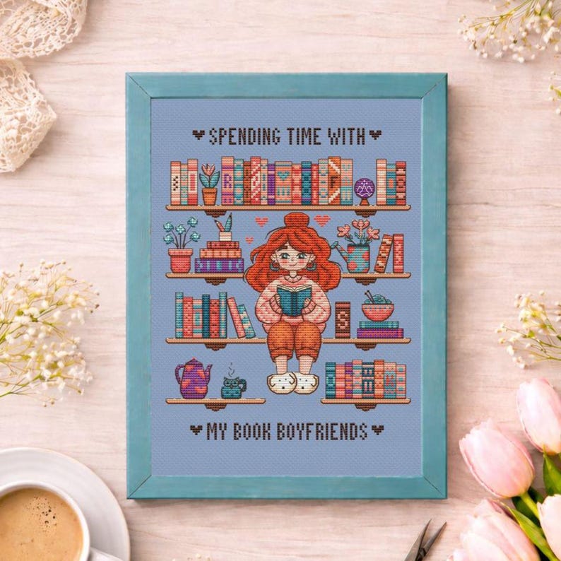 May include: A framed cross-stitch embroidery on a light blue background featuring a person with red hair reading a book. The design includes shelves filled with colorful books, potted plants, a teapot, and the text SPENDING TIME WITH MY BOOK BOYFRIENDS.