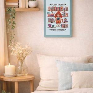 May include: A framed cross-stitch artwork with a light blue frame. The artwork features a cartoon illustration of a person reading, surrounded by bookshelves. Text reads "Spending Time With" and "My Book Boyfriends."