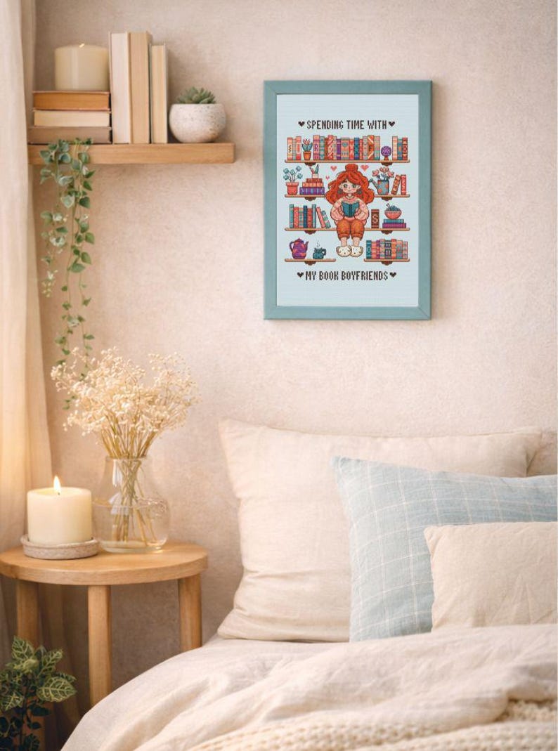 May include: A framed cross-stitch artwork with a light blue frame. The artwork features a cartoon illustration of a person reading, surrounded by bookshelves. Text reads "Spending Time With" and "My Book Boyfriends."