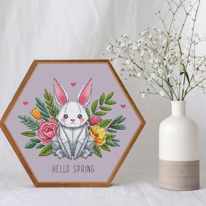 May include: A framed cross-stitch artwork featuring a white rabbit surrounded by colorful flowers and greenery. The artwork is in a hexagonal wooden frame and has the words "HELLO SPRING" at the bottom. A white vase with flowers is next to the artwork.