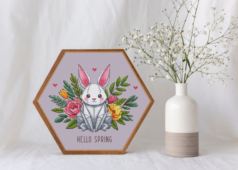 May include: A framed cross-stitch artwork featuring a white rabbit surrounded by colorful flowers and greenery. The artwork is in a hexagonal wooden frame and has the words "HELLO SPRING" at the bottom. A white vase with flowers is next to the artwork.