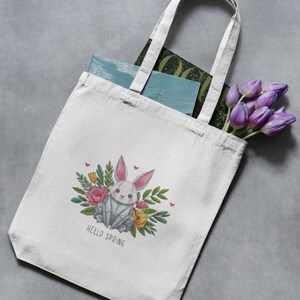 May include: White canvas tote bag featuring a geometric bunny design surrounded by flowers and the text "HELLO SPRING". The bag contains books and a bouquet of purple tulips. The tote bag has two white handles.