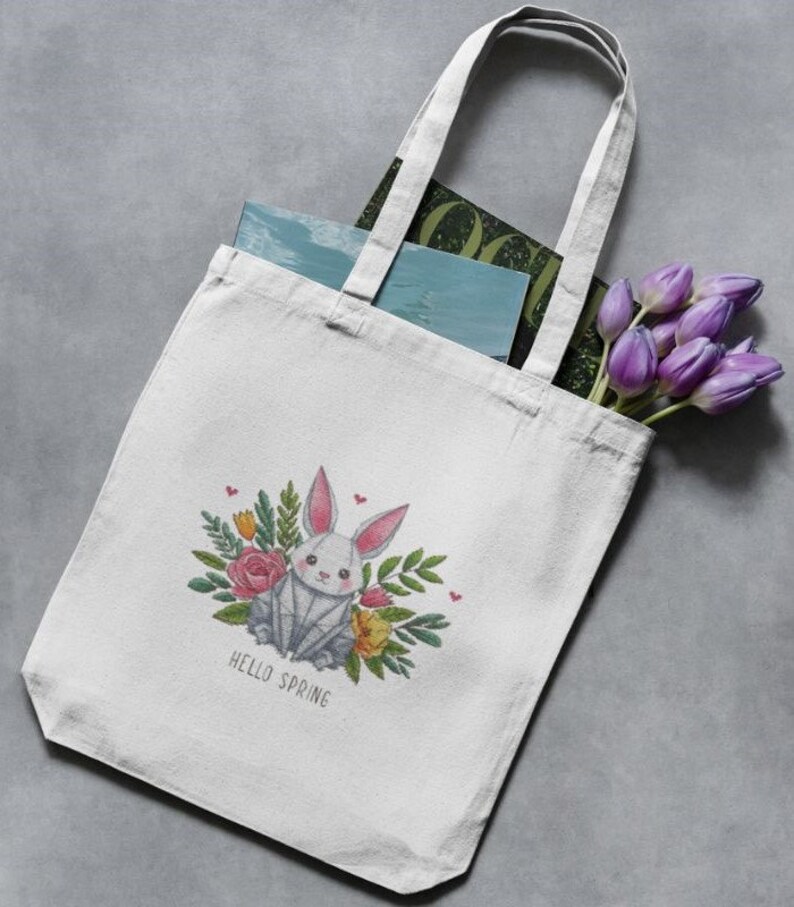 May include: White canvas tote bag featuring a geometric bunny design surrounded by flowers and the text "HELLO SPRING". The bag contains books and a bouquet of purple tulips. The tote bag has two white handles.