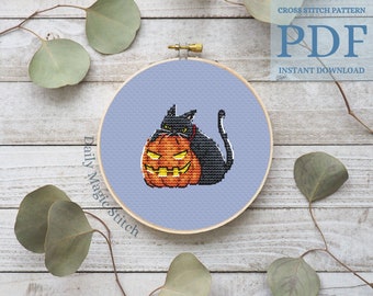 May include: Cross stitch pattern featuring a black cat sitting on a jack-o'-lantern. The pattern is displayed in a wooden embroidery hoop on a light wood background. The text "Daily Magic Stitch" is visible on the hoop.
