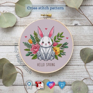 May include: Cross stitch pattern showing a geometric white bunny with pink ears, surrounded by flowers and greenery. The text "HELLO SPRING" is below the bunny. The design is set in a wooden embroidery hoop.