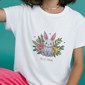 May include: White t-shirt featuring a gray origami-style bunny surrounded by flowers and greenery. The design includes pink, yellow, and green elements, with the text "HELLO SPRING" below the bunny.