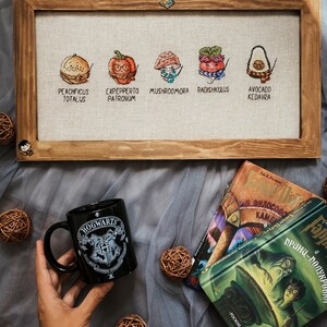 May include: A framed cross-stitch featuring five whimsical food items with Harry Potter-themed names: Peachficus Totalus, Expepperto Patronum, Mushroomora, Radishkulus, and Avocado Kedavra. A black Hogwarts mug and books are also in the image.