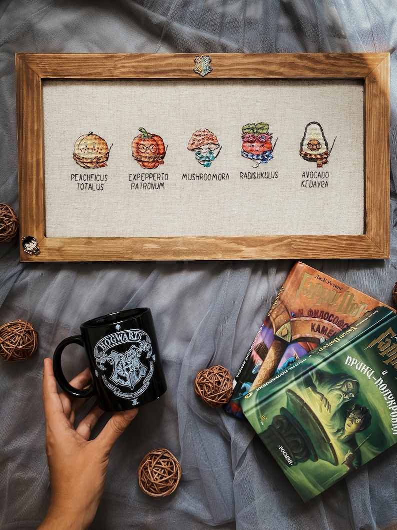 May include: A framed cross-stitch featuring five whimsical food items with Harry Potter-themed names: Peachficus Totalus, Expepperto Patronum, Mushroomora, Radishkulus, and Avocado Kedavra. A black Hogwarts mug and books are also in the image.