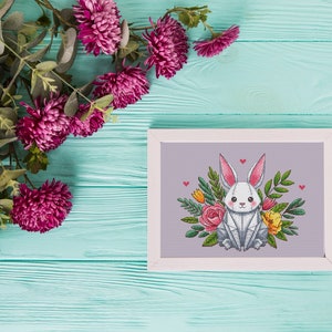 May include: A framed cross-stitch of a white bunny with pink-lined ears, surrounded by flowers and greenery. The artwork is set against a light purple background, with a white frame. Decorative pink flowers and green leaves are in the upper left corner.