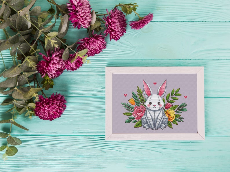 May include: A framed cross-stitch of a white bunny with pink-lined ears, surrounded by flowers and greenery. The artwork is set against a light purple background, with a white frame. Decorative pink flowers and green leaves are in the upper left corner.