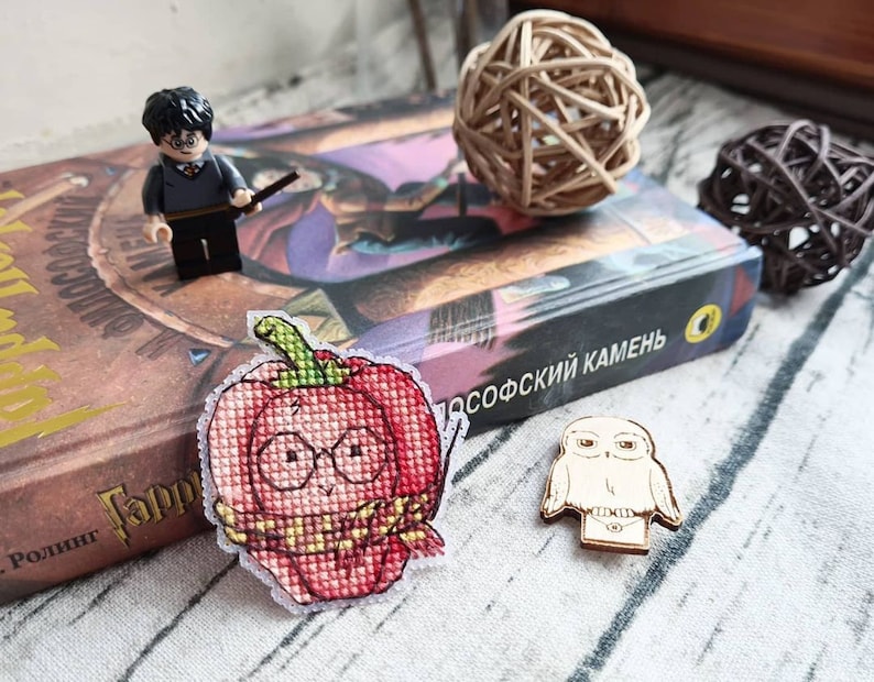 May include: A cross-stitched patch in the shape of a red apple with glasses and a scarf, a wooden owl, and a Lego figure of a boy with a wand are displayed on a book. Decorative wicker balls are also present.