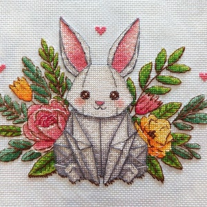 May include: Cross-stitch design of a gray origami-style bunny surrounded by flowers and greenery. The bunny has pink inner ears and rosy cheeks. Pink hearts are scattered around the bunny and flowers. The flowers are pink, orange, and yellow.