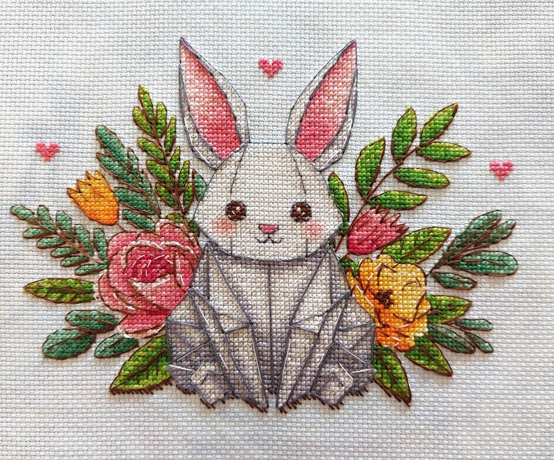 May include: Cross-stitch design of a gray origami-style bunny surrounded by flowers and greenery. The bunny has pink inner ears and rosy cheeks. Pink hearts are scattered around the bunny and flowers. The flowers are pink, orange, and yellow.