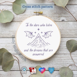 May include: A white embroidery hoop with a black and white line drawing of a mountain with two wings and stars above it. The text "To the stars who listen and the dreams that are answered" is written in black below the mountain. The hoop is on a wooden surface.
