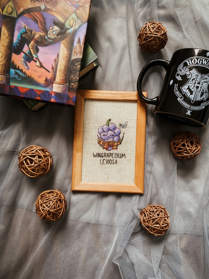 May include: A framed cross-stitch of a purple grape-like design with the text "WINGRAPEDIUM LEVIOSA". A black Hogwarts mug, a Harry Potter book, and decorative woven balls are also present.