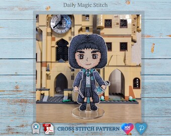 May include: Cross stitch pattern of a cartoon character with black hair, wearing a blue and white uniform, holding a wand. The character is standing in front of a brick building with a clock tower.