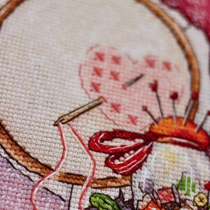 May include: Close-up of a cross-stitch project in progress. The image shows a wooden embroidery hoop holding fabric with a heart design, a needle, and a pin cushion. Red and white thread is visible.