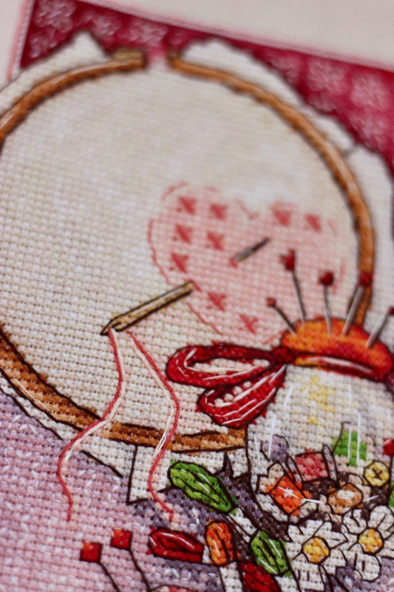 May include: Close-up of a cross-stitch project in progress. The image shows a wooden embroidery hoop holding fabric with a heart design, a needle, and a pin cushion. Red and white thread is visible.