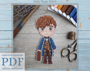 May include: Cross stitch pattern PDF download of a cartoon illustration of a young man with brown hair, wearing a blue coat, brown pants, and a yellow scarf. He is holding a brown suitcase and a wand. The image is on a wooden background with embroidery floss and scissors.