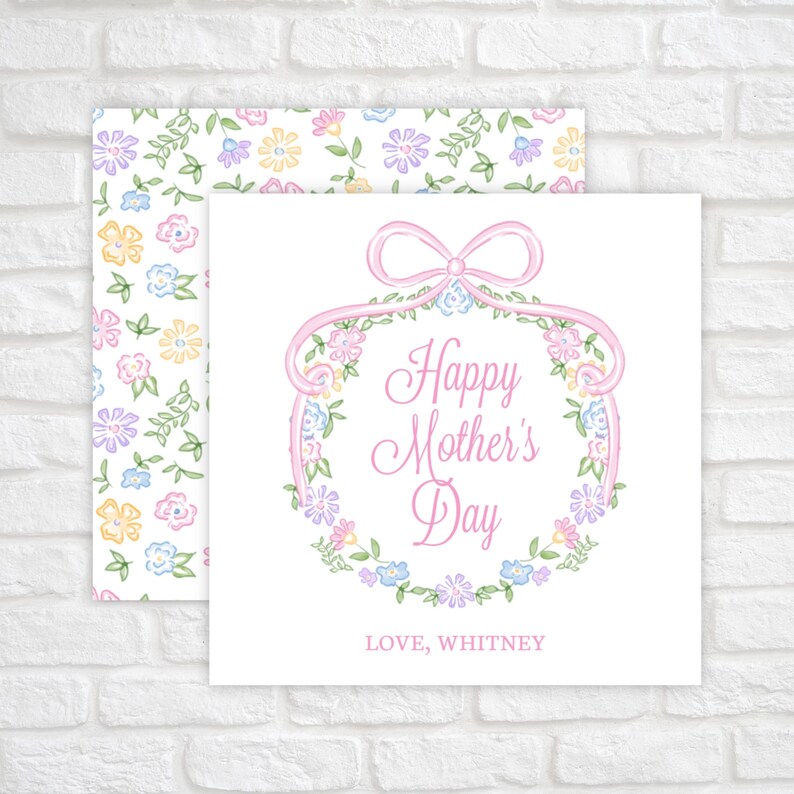 Op de afbeelding: Een witte kaart met een roze lint en een krans van bloemen. De tekst luidt "Happy Mother's Day" en "LIEFD, WHITNEY".