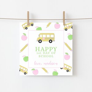 May include: A square card with a school theme, featuring a yellow school bus, apples, and pencils on a white background. The words "HAPPY 1st DAY OF SCHOOL" are in green, with "love, madison" in pink.