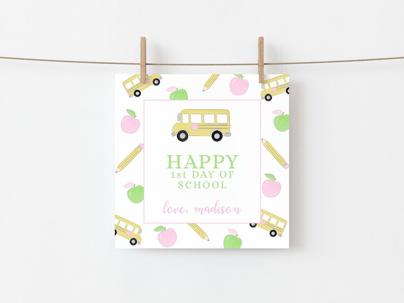 May include: A square card with a school theme, featuring a yellow school bus, apples, and pencils on a white background. The words "HAPPY 1st DAY OF SCHOOL" are in green, with "love, madison" in pink.