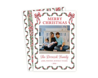 May include: A rectangular Christmas card featuring a central photograph of a family of four. The card has a white border decorated with green holly leaves, red berries, and pink bows. Text reads MERRY CHRISTMAS, The Dronzek Family, JOHN, WHITNEY, HUNTER & TUCKER, and 2026.