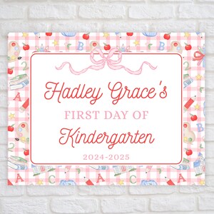 May include: A pink and white gingham patterned sign with a red border. The sign says "Hadley Grace's First Day of Kindergarten 2024-2025". The sign is decorated with a pink bow, apples, pencils, books, and stars.