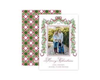 May include: A rectangular Christmas card featuring a central photograph of a family of four, framed by a watercolor illustration of green holly leaves, red berries, and a pink ribbon. The text reads 2025, Merry Christmas, and the names John, Whitney, Hunter, Tucker Dronzek.