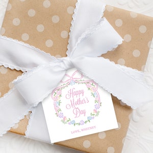 Op de afbeelding: Een wit cadeau-etiket met een roze bloemenkrans en de tekst "Happy Mother's Day" in roze. Het etiket is vastgebonden aan een bruine cadeaudoos met witte stippen met een wit lint.