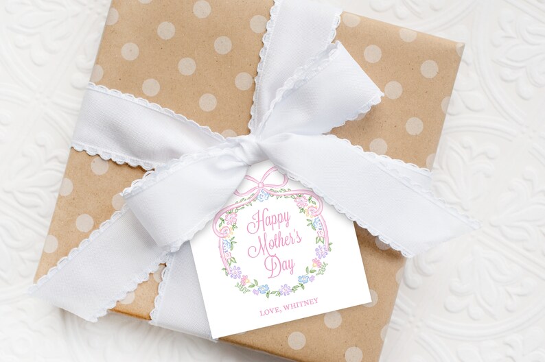 Op de afbeelding: Een wit cadeau-etiket met een roze bloemenkrans en de tekst "Happy Mother's Day" in roze. Het etiket is vastgebonden aan een bruine cadeaudoos met witte stippen met een wit lint.