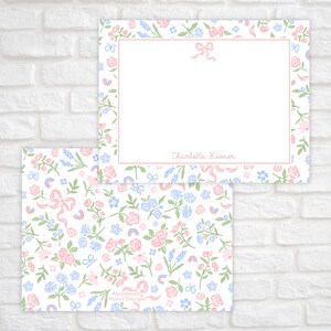 Puede incluir: Una tarjeta blanca con un borde floral rosa y un lazo rosa en la parte superior. La tarjeta tiene un espacio en blanco para escribir y el texto "Charlotte Keener" en la parte inferior. La tarjeta est&aacute; sobre un fondo de ladrillo blanco. La parte posterior de la tarjeta tiene un patr&oacute;n repetitivo de flores rosas y azules, lazos y arco&iacute;ris.