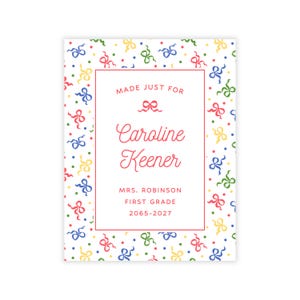May include: A rectangular white sign with a red border featuring a pattern of colorful bows and dots. The text reads: MADE JUST FOR Caroline Keener MRS. ROBINSON FIRST GRADE 2065-2027. The design includes red, blue, yellow, and green elements.