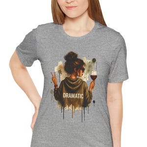 May include: A heather gray t-shirt featuring a watercolor graphic of a person with a bun, sunglasses, and a glass of wine. The word "DRAMATIC" is printed on the hoodie. The person is making a peace sign.