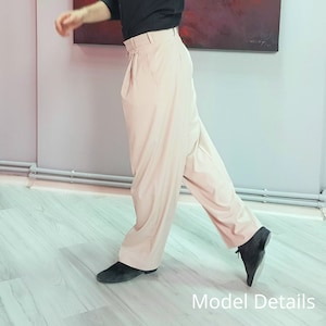 May include: A person wearing a black shirt and beige trousers with pleats. The trousers are wide-legged and the person is standing on a wooden floor.