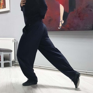Op de afbeelding: Een persoon in een zwarte broek en een zwart shirt staat in een dansstudio. De persoon staat in een danspose met &eacute;&eacute;n been naar voren gestrekt en het andere been gebogen bij de knie. De persoon draagt zwarte schoenen.