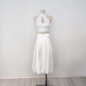 May include: A two-piece formal outfit on a mannequin. The top is a white sequined halter with a keyhole cutout. The skirt is a white, flowing midi-length design with a side slit. The outfit is displayed on a neutral background.