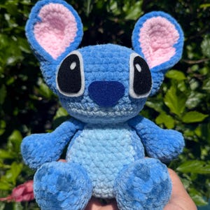 Puede incluir: Un peluche azul hecho a mano con ganchillo, con grandes ojos negros, una nariz azul oscuro y orejas forradas de rosa. El peluche es un personaje de una popular pel&iacute;cula de animaci&oacute;n, con un aspecto suave y texturizado. El juguete se sostiene en una mano.