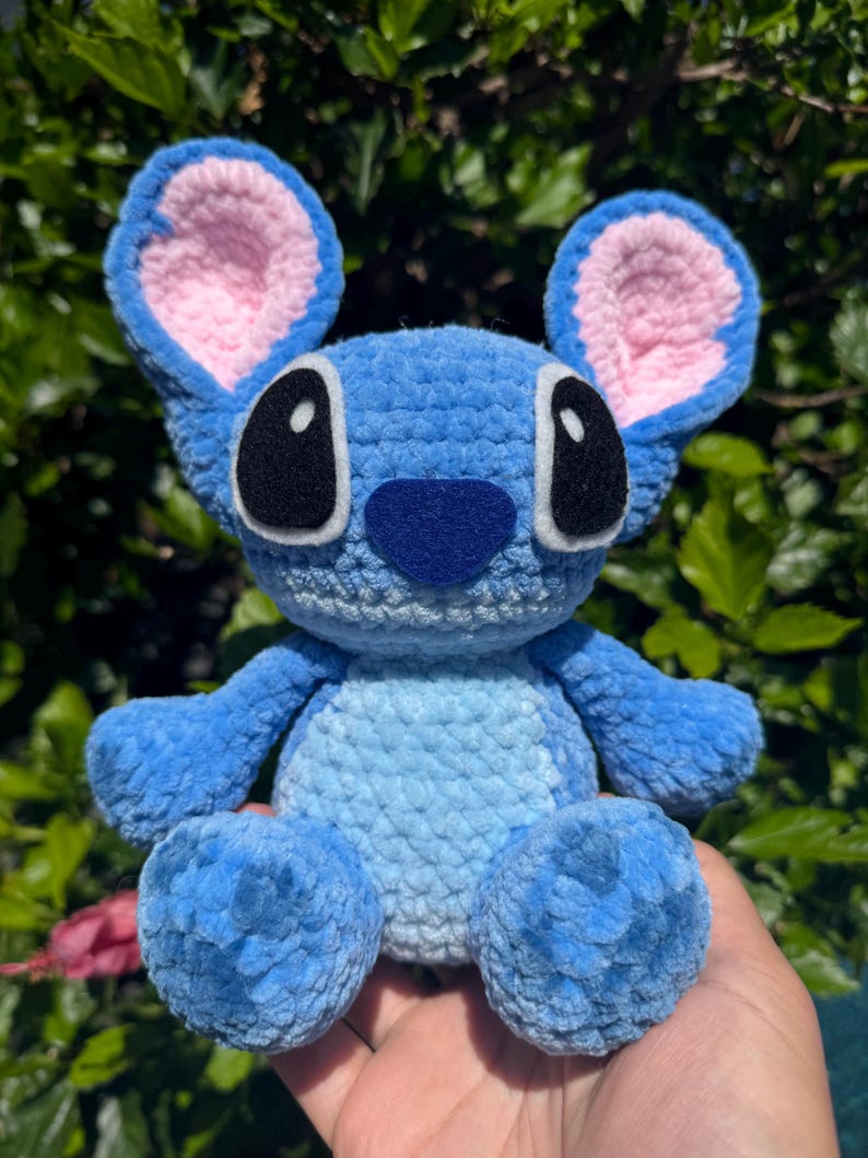 Puede incluir: Un peluche azul hecho a mano con ganchillo, con grandes ojos negros, una nariz azul oscuro y orejas forradas de rosa. El peluche es un personaje de una popular pel&iacute;cula de animaci&oacute;n, con un aspecto suave y texturizado. El juguete se sostiene en una mano.