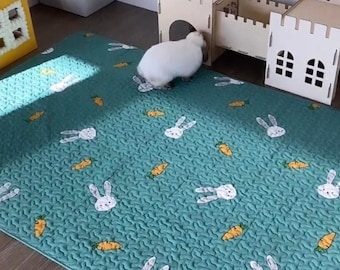 Op de afbeelding: Een turquoise gewatteerde mat met een patroon van witte konijnen en oranje wortels. Een wit konijn staat in de buurt van een houten constructie. De mat is ontworpen voor een huisdier.