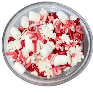 May include: A clear glass bowl containing a mix of red, white, and pink sprinkles. The mix includes red sprinkles, snowflake-shaped sprinkles, and white marshmallows. The sprinkles are a festive mix, ideal for decorating cakes and other treats.