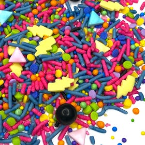 May include: A colorful assortment of sprinkles, including pink, blue, yellow, and green shapes. The mix features rod-shaped sprinkles, small spheres, triangles, and lightning bolt shapes. Two black spheres are also visible.