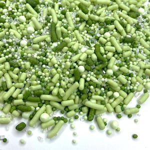 May include: A close-up of a pile of green sprinkles in various shapes and sizes. The sprinkles include small round balls, long rods, and irregular shapes in shades of light and dark green. Some clear, crystalline sugar pieces are also visible.
