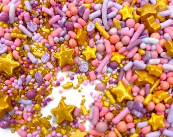 May include: A mix of pink, purple, and gold sprinkles. The sprinkles are in the shape of stars, balls, and small sticks.
